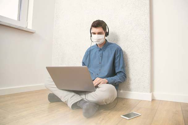 Ένας τύπος με προστατευτική μάσκα δουλεύει σε έναν υπολογιστή σε ένα άδειο δωμάτιο στο σπίτι κατά τη διάρκεια επιδημίας ιού. Καραντίνα κατά τη διάρκεια ιογενούς λοίμωξης - Φωτογραφία, εικόνα