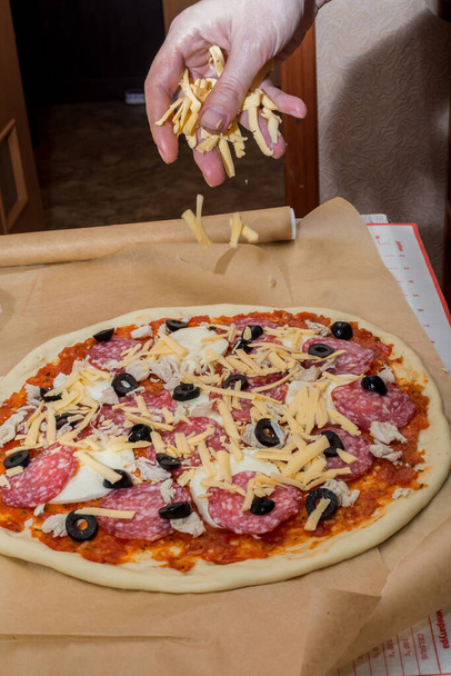 šéfkuchař syrovou pizzu posype strouhaným sýrem. Dělám pizzu. syrové těsto na pečicím papíru. omáčka z rajčat, cibule, česneku, oregána a bylin, mozzarelly a černých oliv. Vařit doma. uvaříme si sami. - Fotografie, Obrázek