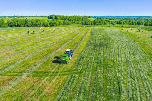 Maataloustraktori kerää niitettyä ruohoa maatalouskäyttöön ja käärii heinäpaaleja muovipellolle, maaseudulle maisema-antenninäköala - Valokuva, kuva