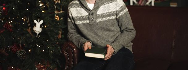 Bir adam Noel ağacının yanındaki bir kazakla kanepede oturuyor. Bir adam elinde bir kitap tutuyor. Bir kitap okuyarak dinlen.. - Fotoğraf, Görsel