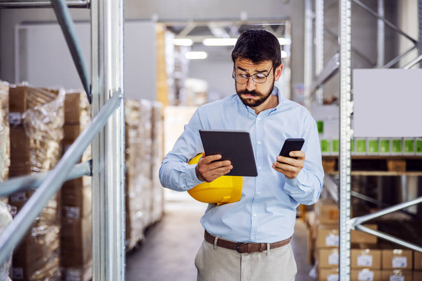 Young handsome bearded employee holding tablet and smart phone in hands and trying to finish work on time. Warehouse interior. - Foto, immagini