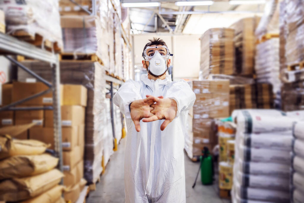Jongeman in beschermende steriele pak en masker staat in het magazijn en bereidt zich voor om het interieur te steriliseren van corona virus / covid-19. - Foto, afbeelding