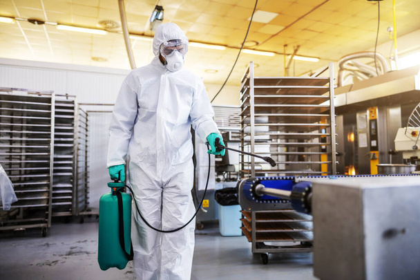 Hombre con traje protector y máscara desinfectando la fábrica de alimentos llena de productos alimenticios del virus corona / covid-19
. - Foto, imagen