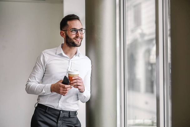 Молодой улыбающийся красивый элегантный бизнесмен опирается на колонну, держа в руках смартфон и чашку свежего утреннего кофе во время кофе-брейка
. - Фото, изображение