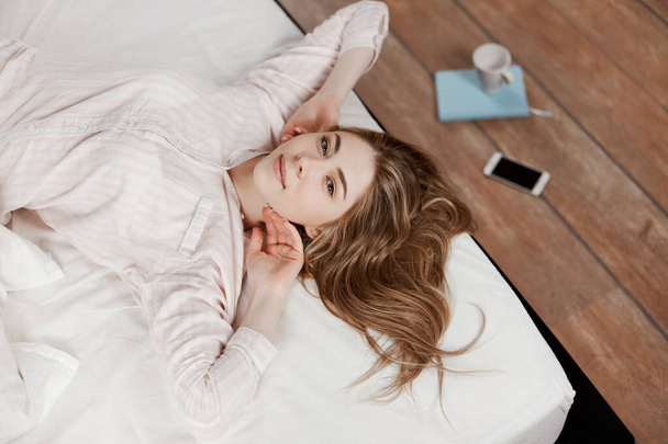Una chica gentil se estira en la cama después de despertarse y sonríe, al lado del suelo hay un Bloc de notas, teléfono y una taza de café. La vista desde arriba, lugar para el texto
 - Foto, Imagen
