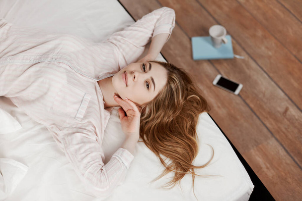 Una chica gentil se estira en la cama después de despertarse y sonríe, al lado del suelo hay un Bloc de notas, teléfono y una taza de café. La vista desde arriba, lugar para el texto
 - Foto, imagen