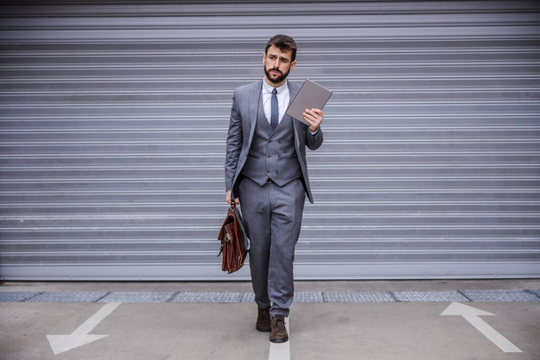 Longitud completa de hombre de negocios guapo caminando hacia la cámara y sosteniendo la tableta en una mano y el maletín en otro
. - Foto, imagen