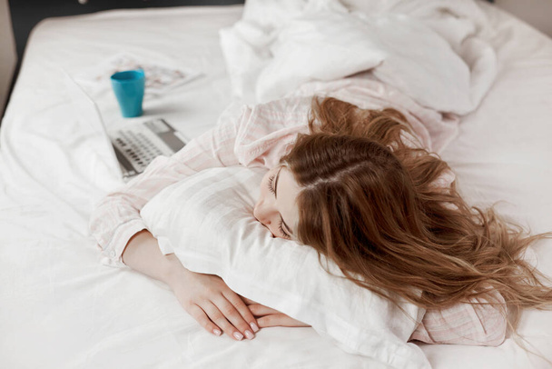 Een jong aantrekkelijk meisje in pyjama ligt thuis op bed, naast een laptop en een kop koffie. Het meisje werkt voor het naar bed gaan - Foto, afbeelding