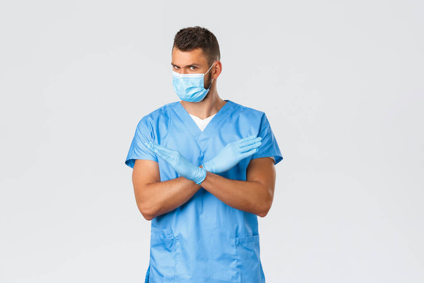 Sağlık çalışanları, covid-19, koronavirüs ve virüs konsepti. Ciddi profesyonel doktor, mavi önlüklü ve tıbbi maskeli erkek hemşire dur diyor, çapraz yasaklama işareti gösteriyor, hoşnutsuz. - Fotoğraf, Görsel
