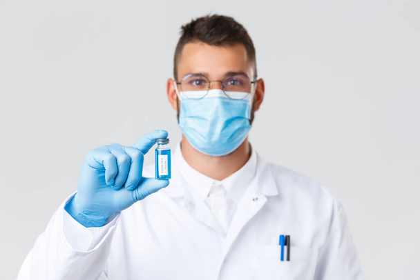 Covid-19, healthcare workers, pandemic and preventing virus concept. Smiling hispanic doctor, physician in medical mask and rubber gloves, showing ampoule with coronavirus vaccine, tell to vaccinate - Фото, изображение
