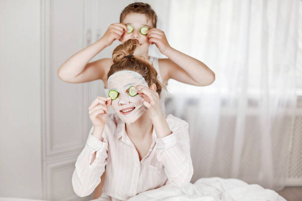 Güzel bir genç kız ve neşeli erkek kardeşi salatalık dilimlerinden yüz maskesi yapıyorlar. Erkek ve kız kardeş gözlerinde salatalıkla yatakta oturuyorlar. Güzellik ve sağlık kavramı - Fotoğraf, Görsel
