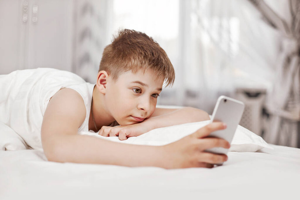 Cute teen boy with brown hair is lying on the bed and looking at the phone. The boy holds the phone in his hand and watches the video - Fotó, kép