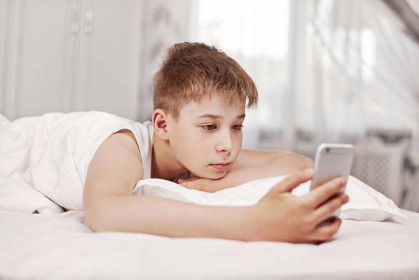 Netter Teenie-Junge mit braunen Haaren liegt auf dem Bett und schaut auf das Telefon. Der Junge hält das Telefon in der Hand und schaut sich das Video an - Foto, Bild