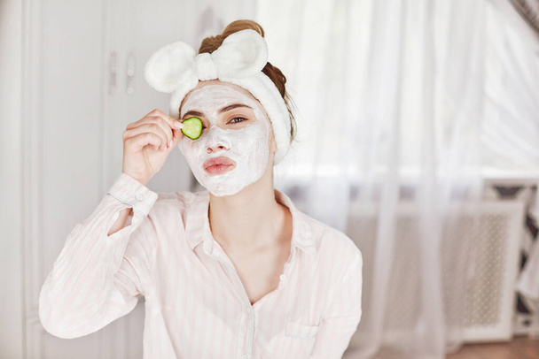 Ein junges Mädchen mit Maske im Gesicht schließt ein Auge mit einer Gurkenscheibe und lächelt. Hautpflege, gesunder Lebensstil. Das Konzept von Schönheit und Gesundheit - Foto, Bild