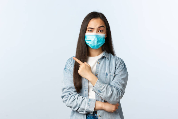 Social distancing lifestyle, covid-19 pandemic everyday life concept. Skeptical displeased asian pretty girl asking explanation, pointing finger left, raise one eyebrow suspicious, wear mask - Photo, image