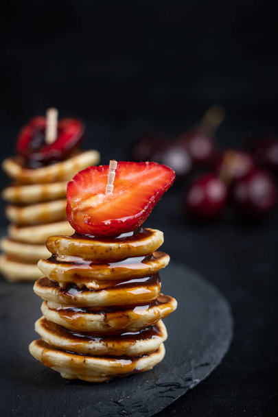 Mini pancakes laying in a stack with strawberry and caramel syrup topping on stony serving platter against dark background - Valokuva, kuva