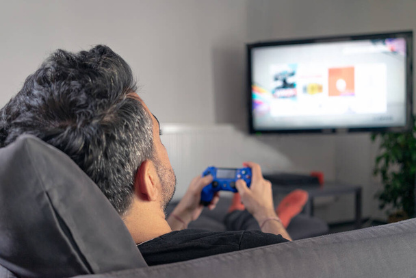 man playing video game at home sitting on coach sofa in front of television with play game concole and joystick in hands - Photo, image