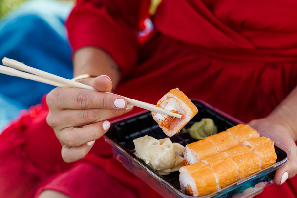 Sushi-Set in Großaufnahme. Attraktives Mädchen im roten Kleid hält Sushi mit Essstäbchen in der Hand. Kurier lieferte Sushi-Set für hübsche Frau. Black Box mit Sushi, Wasabi, Ingwer. - Foto, Bild
