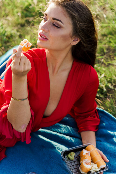 Sushi neben den Lippen eines Mädchens mit blauen Augen beim Picknick. Essenslieferung aus dem japanischen Restaurant. Vertikale Fotowerbung für soziale Netzwerke. Hübsches Mädchen isst Sushi im Park. - Foto, Bild