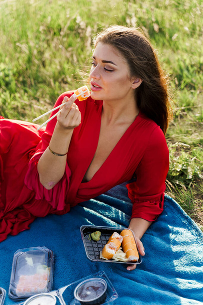 Sushi u úst dívky s modrýma očima na pikniku. Donáška jídla z japonské restaurace. Vertikální foto reklama na sociální sítě. Hezká dívka jí sushi set v parku. - Fotografie, Obrázek