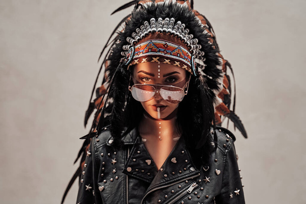Odważna młoda kobieta z azteckim nakryciem głowy, makijażem plemiennym i skórzaną kurtką, pozująca w studio - Zdjęcie, obraz