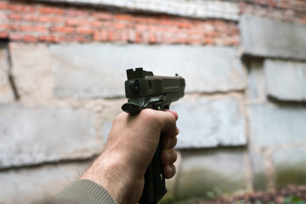 Пистолет в руке кавказца у кирпичной стены заброшенного здания. Стрелок с пистолетом темно-зеленого цвета. Вид от первого лица, пов
. - Фото, изображение
