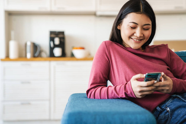 Smiling young asian woman using mobile phone while sitting on a couch at home - Фото, зображення