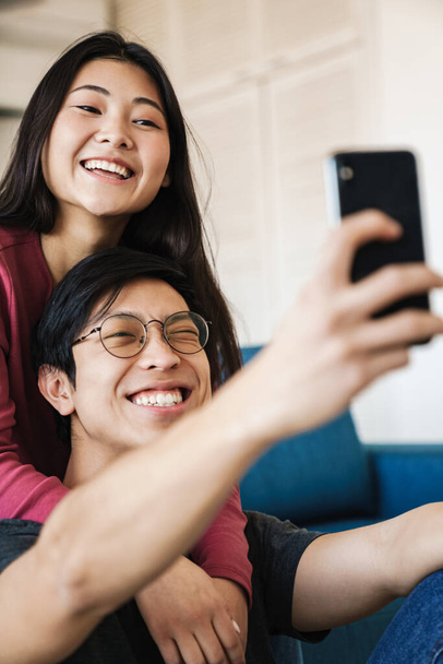 Photo of cheerful asian couple winking and taking selfie photo on mobile phone while sitting at cozy room - Foto, Bild