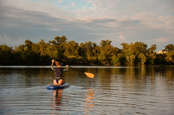 Rear view of woman which sits on sup board holds paddle and floats on the river - Foto, afbeelding