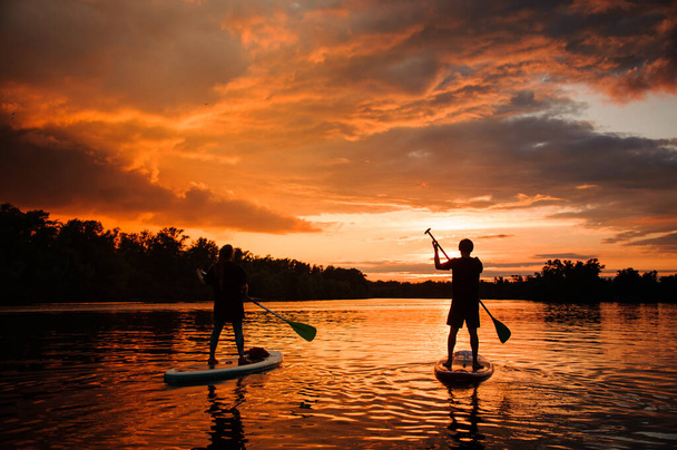 Mooie achteraanzicht van twee personen op sup boards die drijven op de rivier bij zonsondergang - Foto, afbeelding
