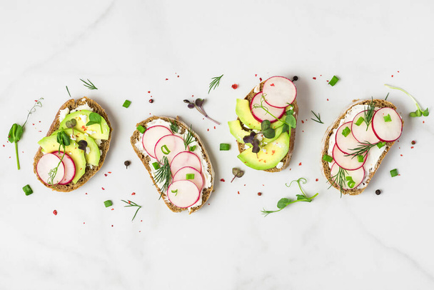 Здорове весняне авокадо та редькові тости з сиром, паростками гороху та перцем на білому мармуровому столі для здорового сніданку. вид зверху
 - Фото, зображення