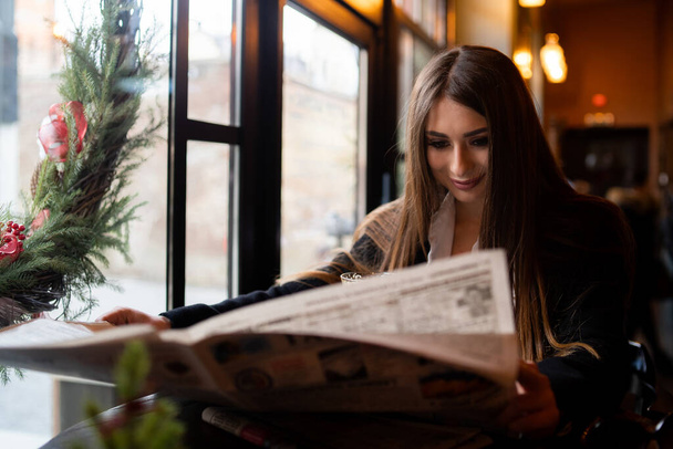 Beautiful woman with brunette hair in dark clothes looking at window and drinking orange hot drink. Concept of cozy places and cold winter. Concept of cafe and coffee shop. - Valokuva, kuva