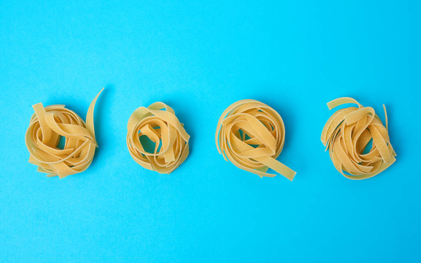gedrehte Pasta in runden Fettuccine-Nestern auf blauem Hintergrund, kreativer Hintergrund mit Lebensmitteln, flache Lage - Foto, Bild