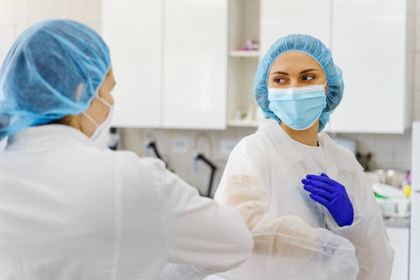 Twee vrouwen met beschermende uitrusting bouffant mob cap en beschermend masker - Elleboog bump begroeting in het ziekenhuis of laboratorium nieuwe normaal als gevolg van corona virus pandemie verspreiding - ondersteuning en veiligheid concept - Foto, afbeelding