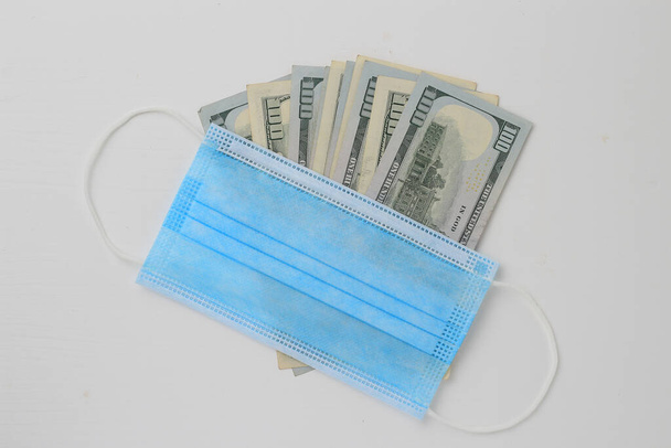 US dollars covered by a classic medical surgical mask on a white wooden background. The crisis is due to the Covid-19 theme - Φωτογραφία, εικόνα