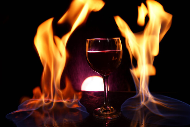 glass of red wine on glass table with fire around it on black background, romantic photo - Фото, зображення