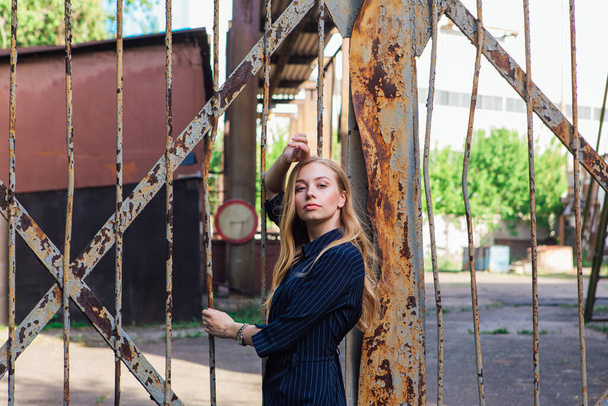 Fashion look's woman with long blond hair standing near the old rusty gate. Young woman modern portrait. - Фото, изображение