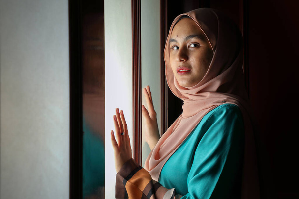 young Asian Malay muslim woman wearing headscarf baju kurung dress at home in front window mood light on face dark background peaceful content calm tranquil break trough wonder - Photo, Image