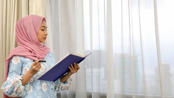 Ev ofisinde başörtüsü takan genç Asyalı Müslüman kadın pencere kenarındaki kitap belgelerine bakıyor. - Fotoğraf, Görsel