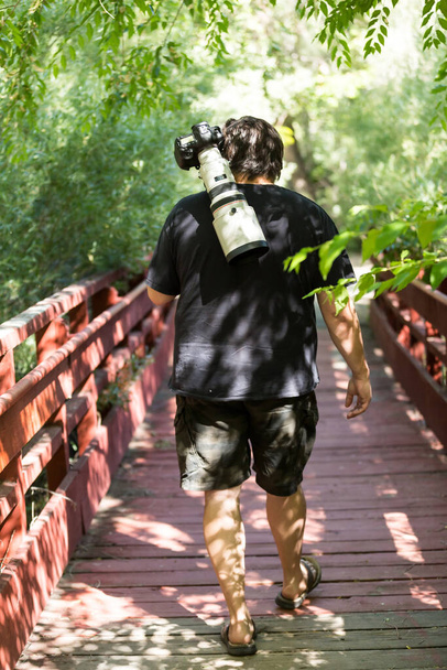 Fotógrafo con lente blanca grande en monópodo caminando en puente buscando vida silvestre - Foto, imagen
