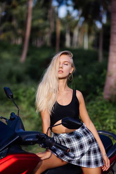 Portré fiatal európai kaukázusi fit vékony nő fekete top fürdőruhában és kockás fekete-fehér iskola lány szoknya trópusi dzsungel területén motorkerékpár, motorkerékpár - Fotó, kép