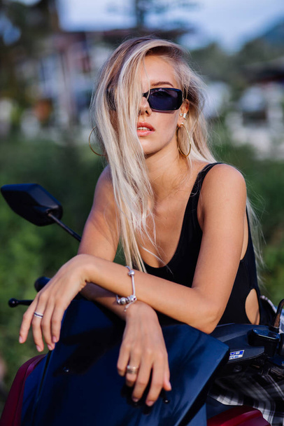 Portrait of young european caucasian fit slim woman in black top swimsuit and plaid black and white school girl skirt on tropical jungle field with motorbike, motorcycle - Фото, зображення