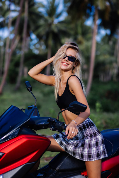 Portrait of young european caucasian fit slim woman in black top swimsuit and plaid black and white school girl skirt on tropical jungle field with motorbike, motorcycle - Fotó, kép
