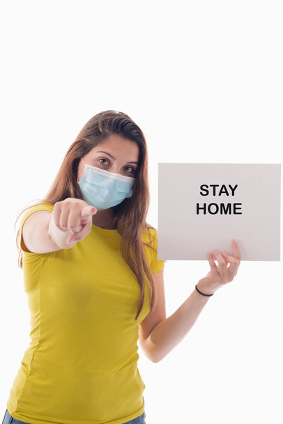 Young female using a surgical mask holding a sign with a message and pointing at the camera on a light background. Safety and awareness concept. - Photo, Image