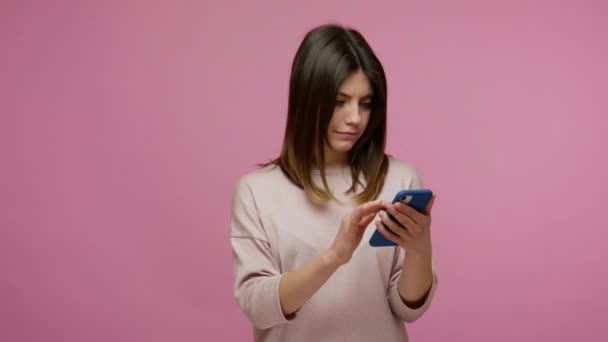 Lovely brunette young woman scrolling internet blog on smartphone, watching photos on social network and expressing amazement, using mobile application. indoor studio shot isolated on pink background - Imágenes, Vídeo