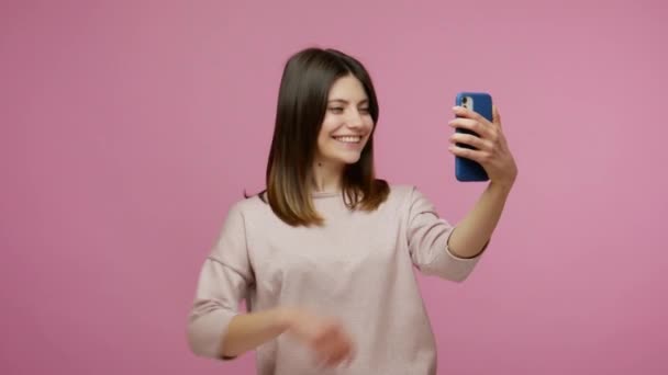Beautiful young brunette woman smiling, having fun while taking selfie on smartphone, using mobile app, making video for social network post, blogging. indoor studio shot isolated on pink background - Filmagem, Vídeo