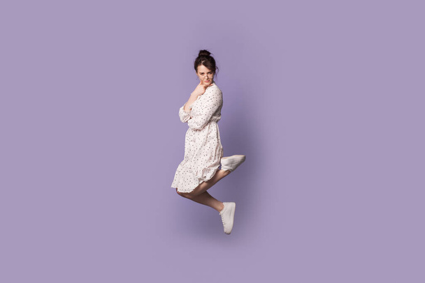 Confused caucasian woman in a summer dress jumping on a violet studio wall and looking at camera - Photo, Image