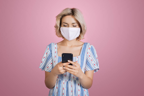 Lovely blonde woman in medical mask wearing a nice dress is chatting on mobile posing on a pink studio wall - Foto, afbeelding