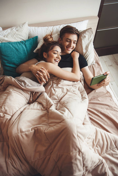 Прекрасная кавказская пара лежит в постели и смотрит что-то в мобиле
 - Фото, изображение