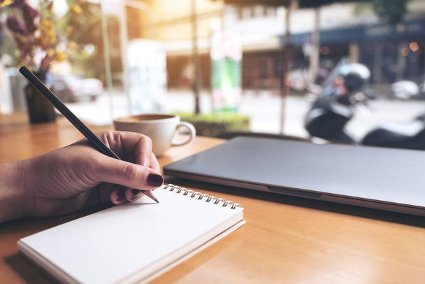 Κοντινό πλάνο εικόνα της γραφής χέρι επιχειρηματία σε ένα κενό σημειωματάριο με φορητό υπολογιστή και φλιτζάνι καφέ σε ξύλινο τραπέζι φόντο - Φωτογραφία, εικόνα